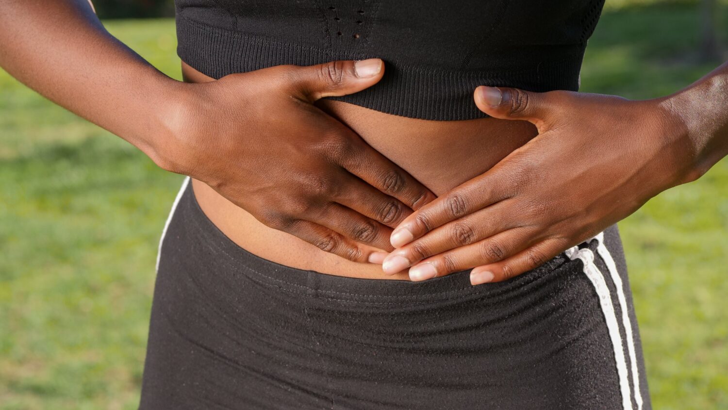 debloat, Woman wearing black sports bra and black workout leggings presses her hands on her stomach.